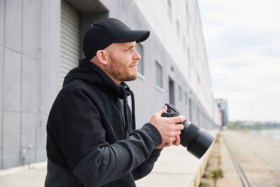Arne Claußen mit schwarzer Kappe und Kapuzenpullover hält eine Kamera und schaut in die Ferne, im Hintergrund ein modernes Gebäude und Bahngleise.