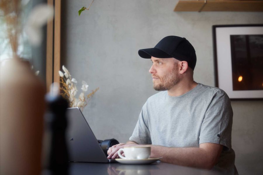 Designer Arne Claußen in grauem T-Shirt und schwarzem Basecap sitzt an einem Tisch in einem Café mit einem Laptop und einer Kaffeetasse, schaut aus dem Fenster.