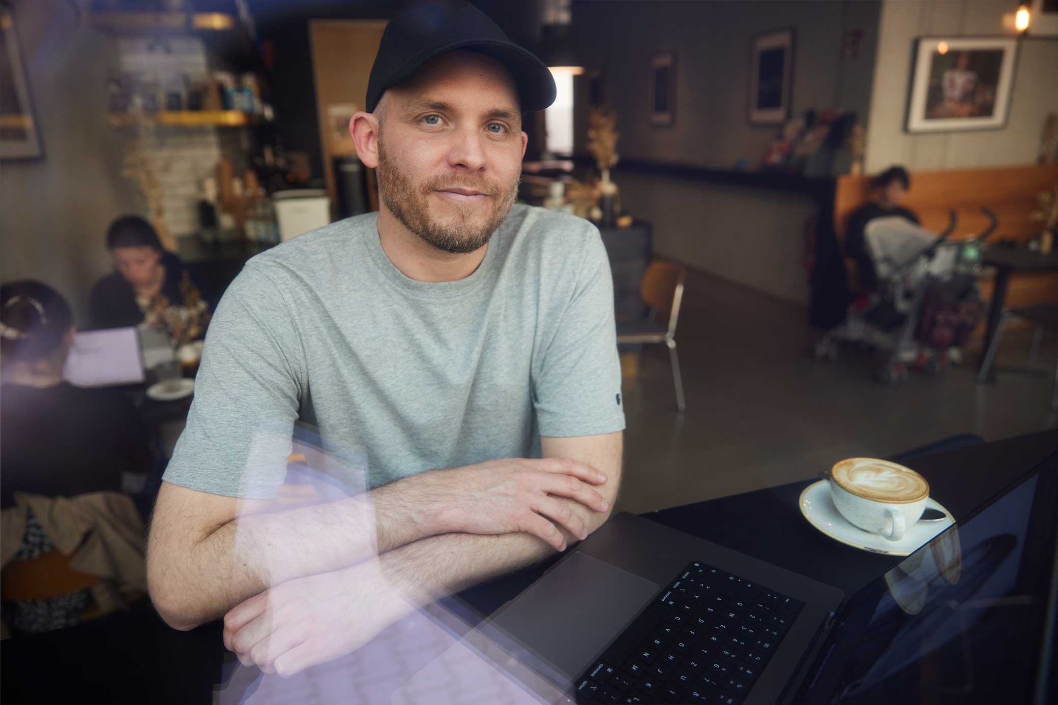 Arne Claußen mit Baseballkappe und grauem T-Shirt sitzt in einem Café vor einem Laptop mit einer Tasse Cappuccino.