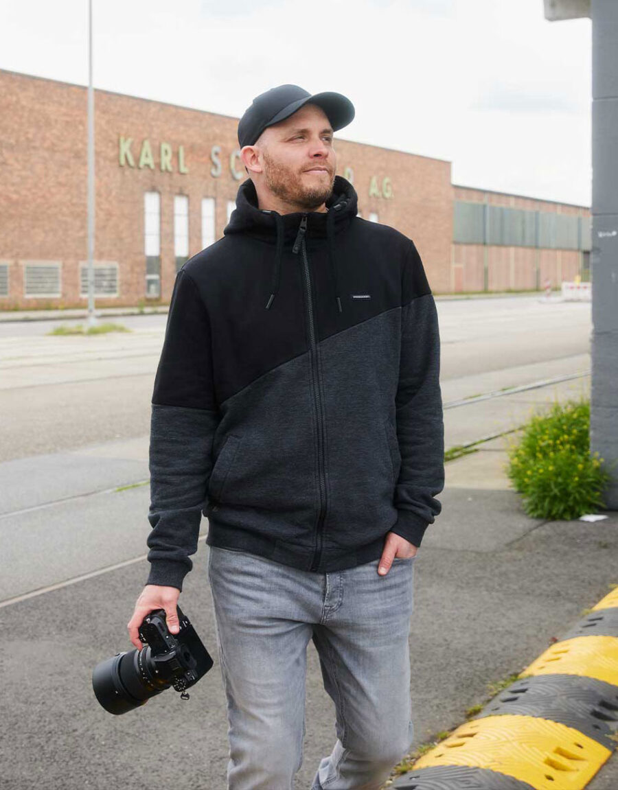 Arne Claußen in grauer Jeans und schwarzem Hoodie hält eine Kamera, steht draußen vor einem Backsteingebäude.