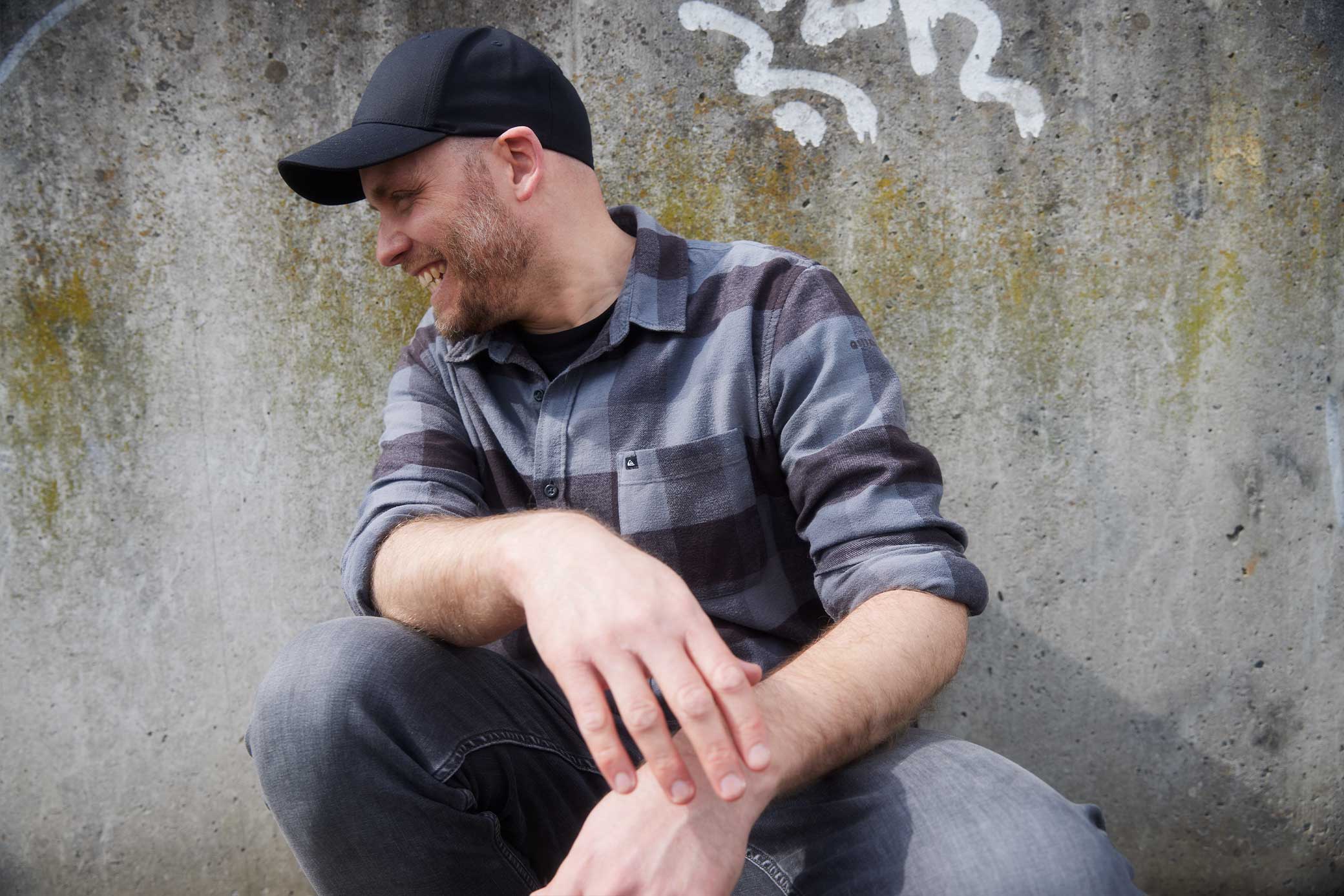 Arne Claußen mit schwarzer Baseballkappe und kariertem Hemd sitzt lächelnd vor einer Betonwand mit Graffitimalereien.