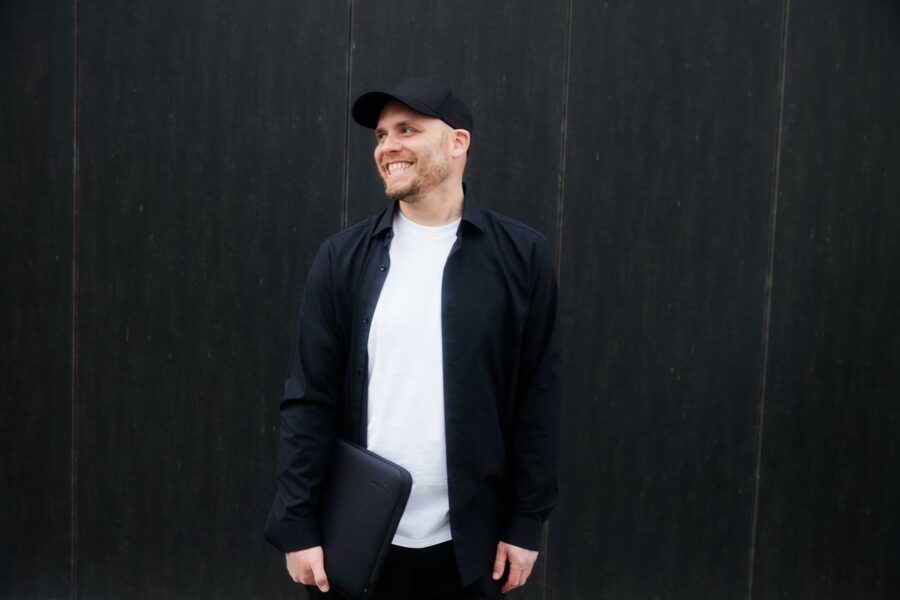 Arne Claußen mit schwarzer Kappe und schwarzem Hemd, lächelt und hält eine schwarze Tasche.
