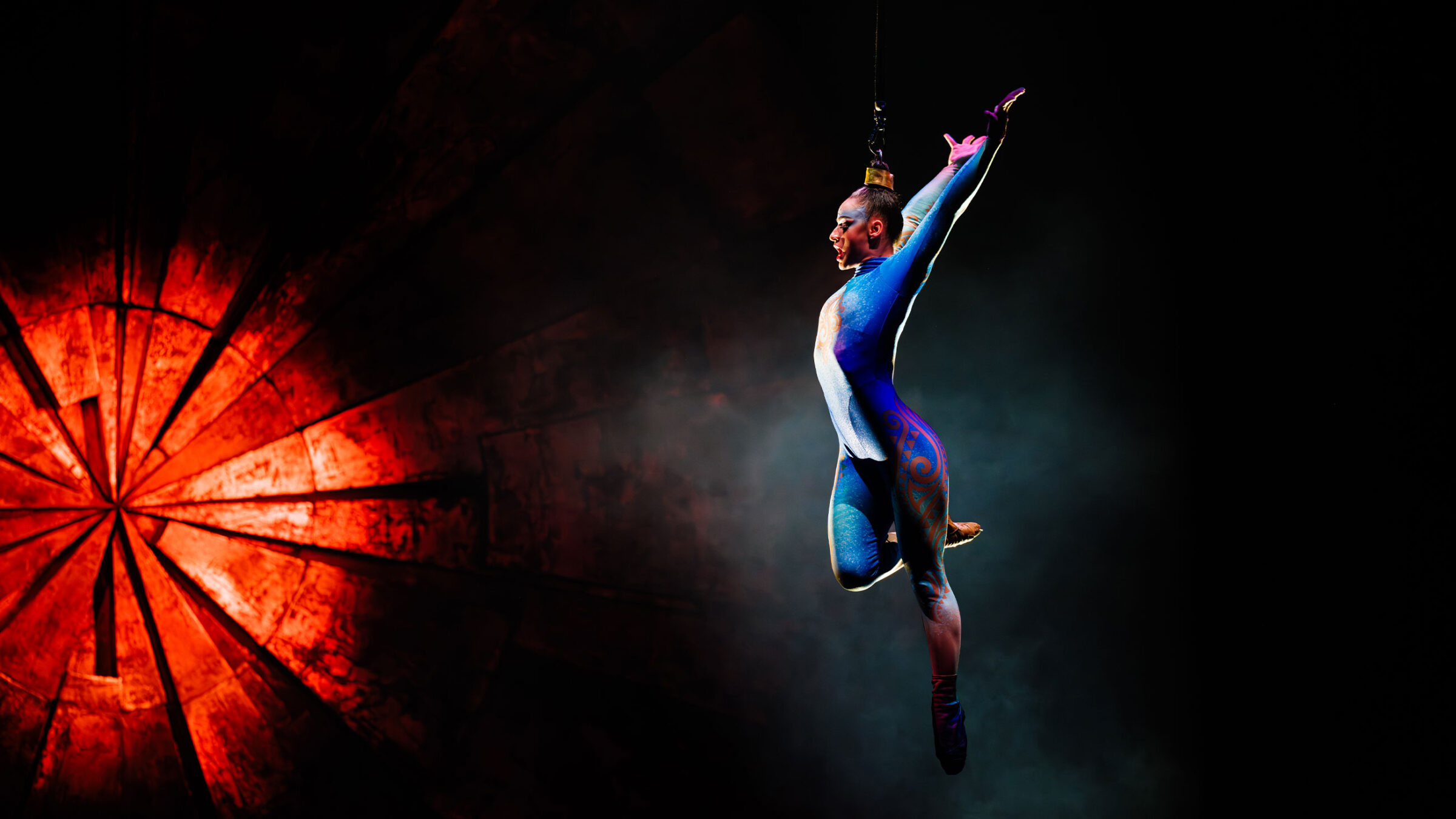 Artistin in blauem Akrobatikanzug führt an einem Seil in der Luft eine Pose aus, vor einem roten und schwarzen Hintergrund.