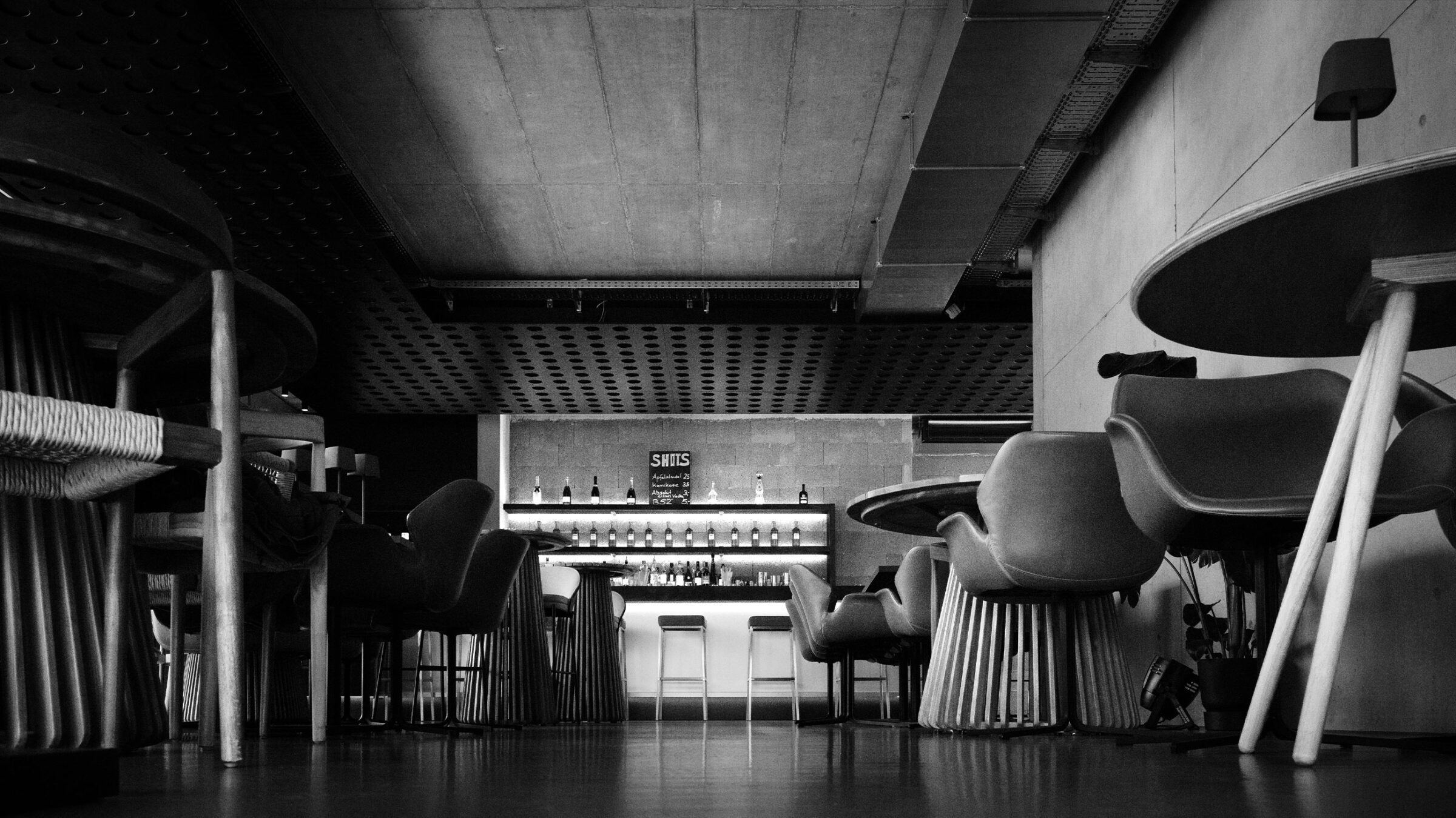 Schwarz-weiß Foto einer modernen Bar mit eleganten Stühlen und Tischen im Vordergrund und einer gut ausgestatteten Theke im Hintergrund.