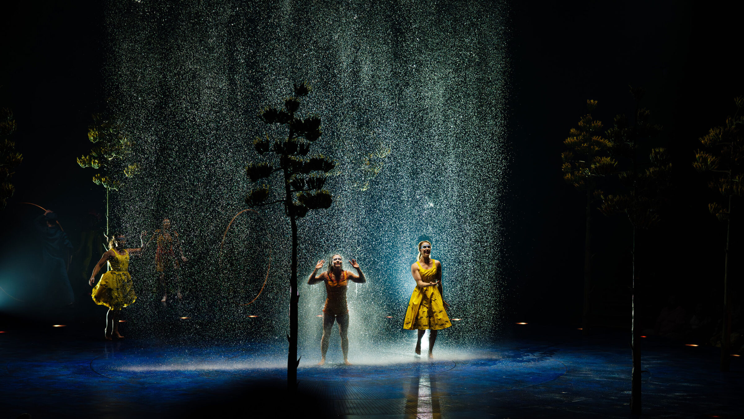 Tänzer in gelben Kostümen performen unter einem Wasserfall auf einer dunklen Bühne. Eine Show von Cirque du Soleil.