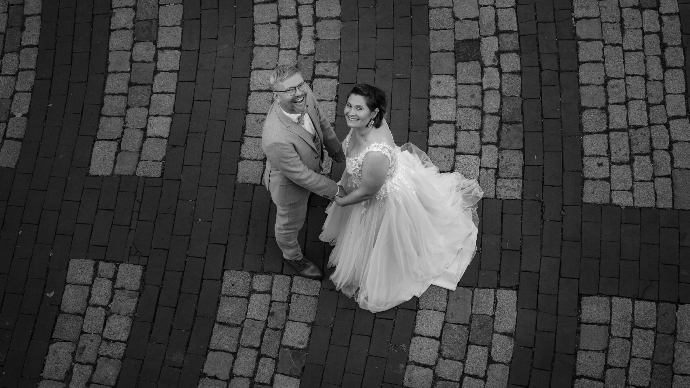Brautpaar in Hochzeitskleidung steht auf einem gepflasterten Weg und lächelt in die Kamera.