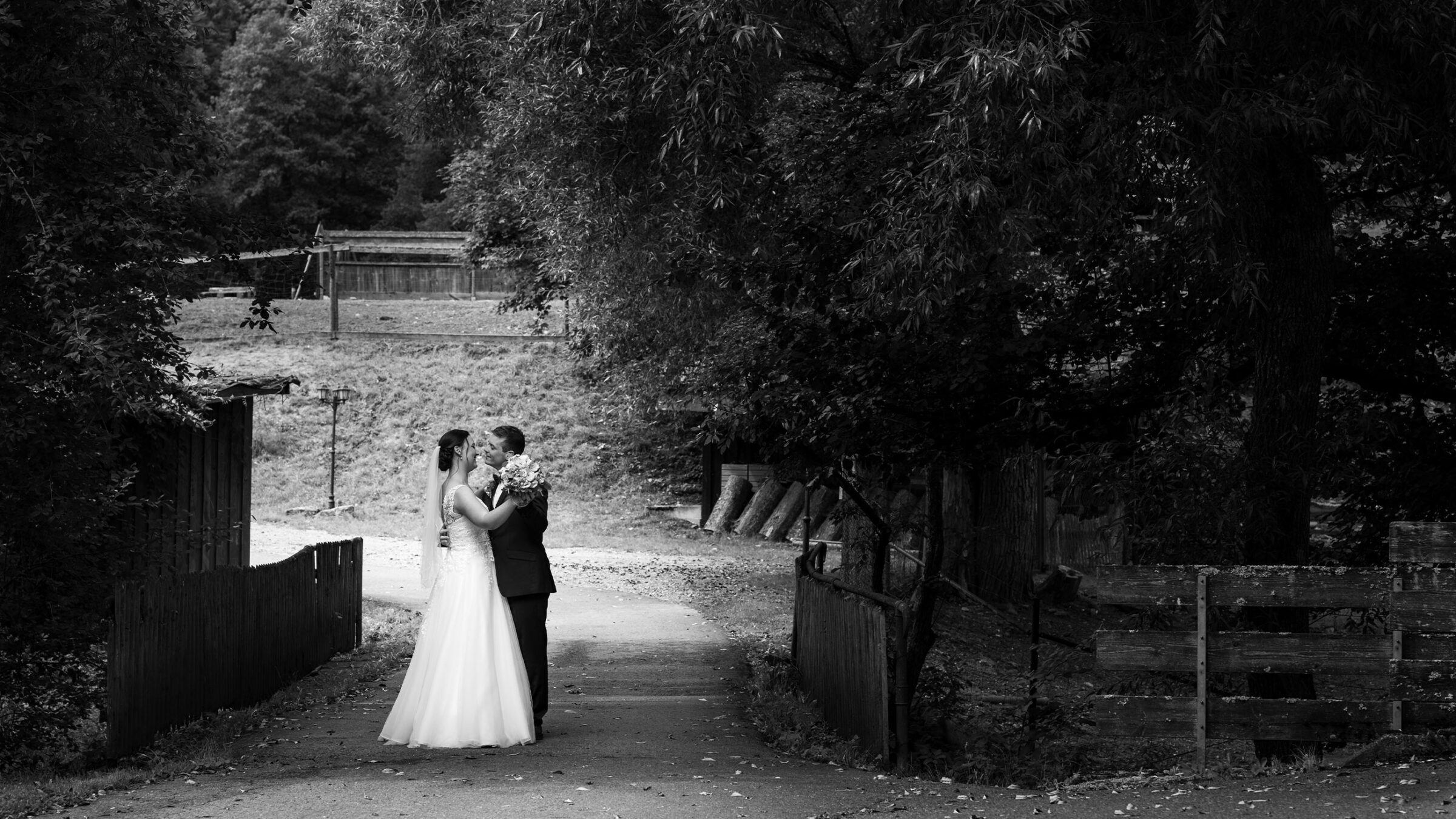 Braut und Bräutigam küssen sich auf einem ländlichen Weg, umgeben von Bäumen und Holzzäunen.