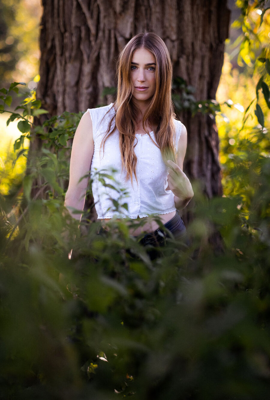 Lea Toran Jenner mit langen, braunen Haaren und weißem ärmellosen Oberteil steht vor einem Baum.