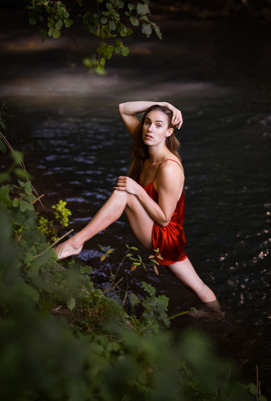 Lea Toran Jenner in einem roten Kleid steht am Rand eines Flusses und schaut in die Kamera, umgeben von grünen Pflanzen.