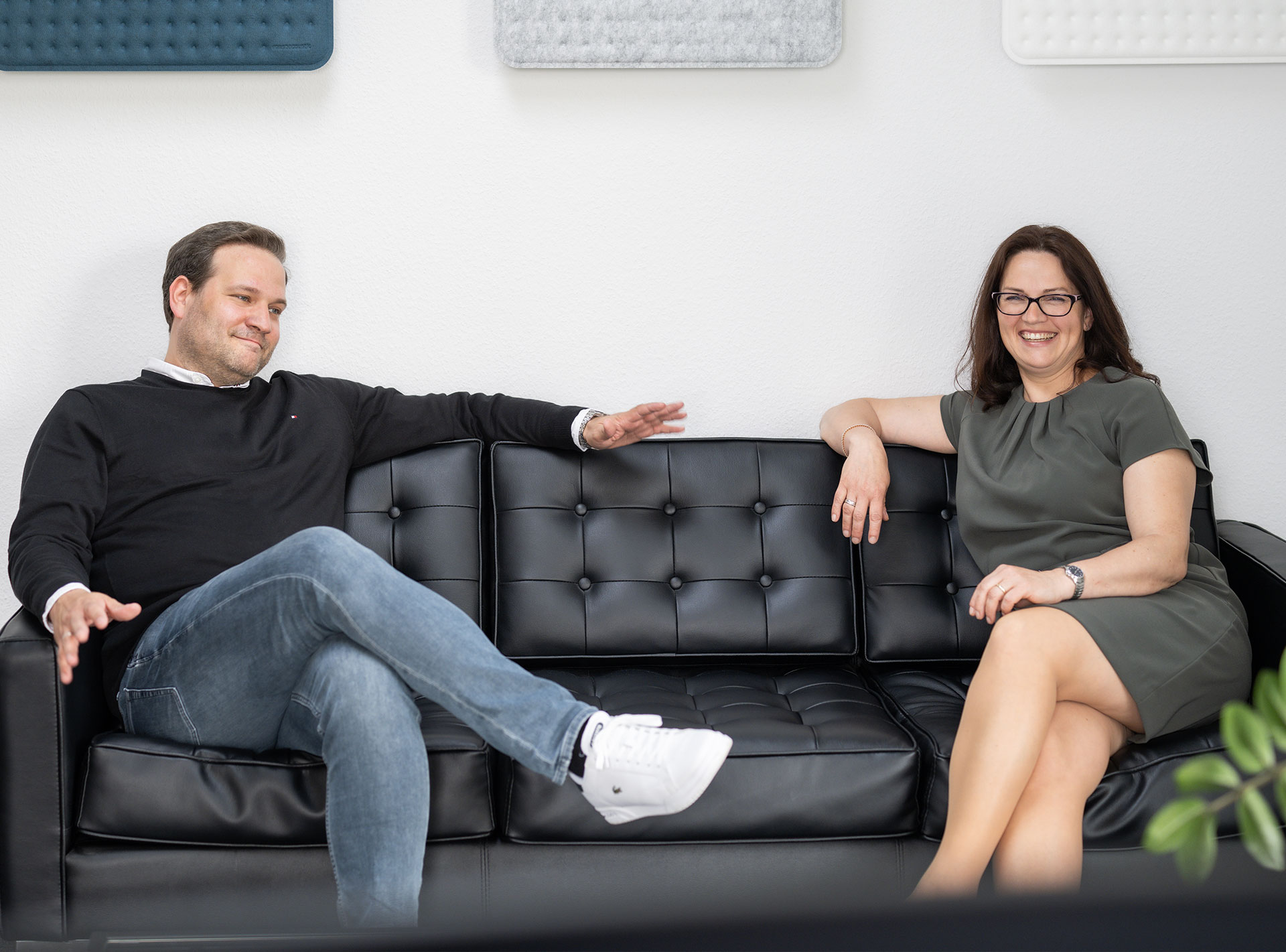Ein Mann und eine Frau sitzen lächelnd auf einem schwarzen Ledersofa in einem modernen Büro.