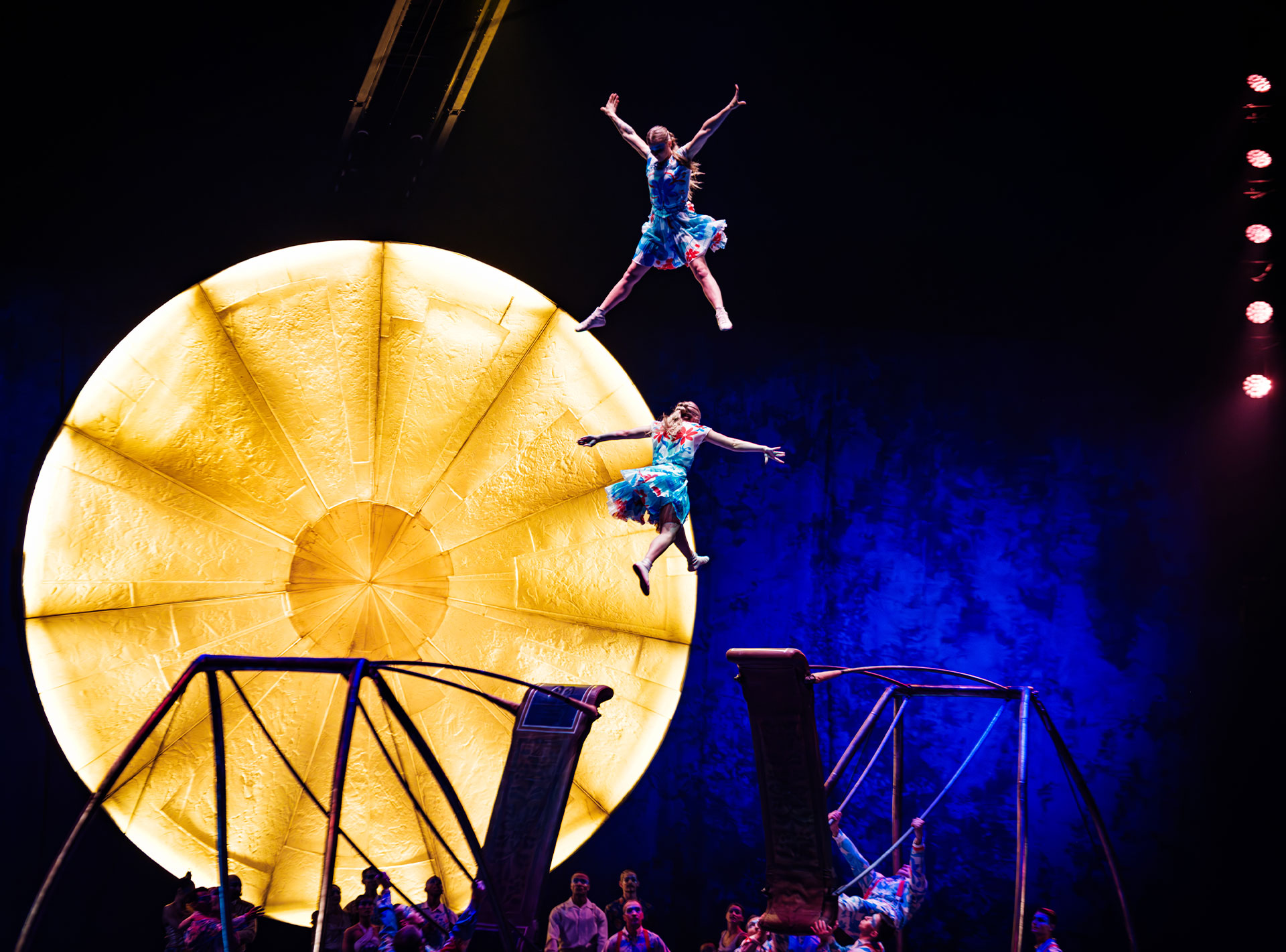 Zwei Akrobaten in bunten Kostümen schweben in der Luft vor einem großen, leuchtenden gelben Kreis auf einer Bühne.