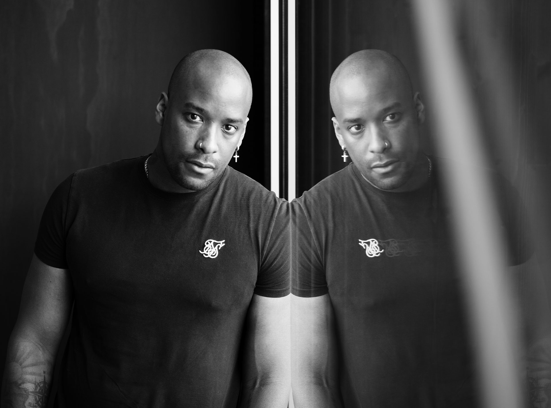 Schwarz-weiß Porträt eines Mannes mit Glatze, der sich an eine Fensterscheibe lehnt und sein Spiegelbild betrachtet.