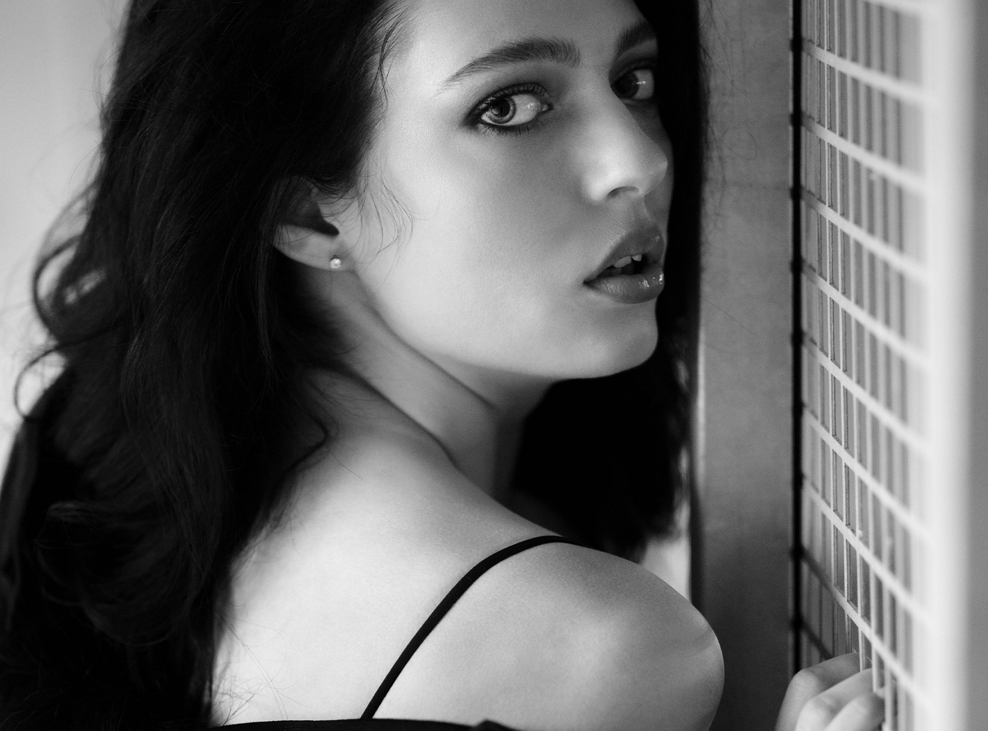 Schwarz-Weiß-Foto einer Frau mit langem dunklem Haar, die an eine Wand mit einem Gittermuster gelehnt ist und über die Schulter blickt.