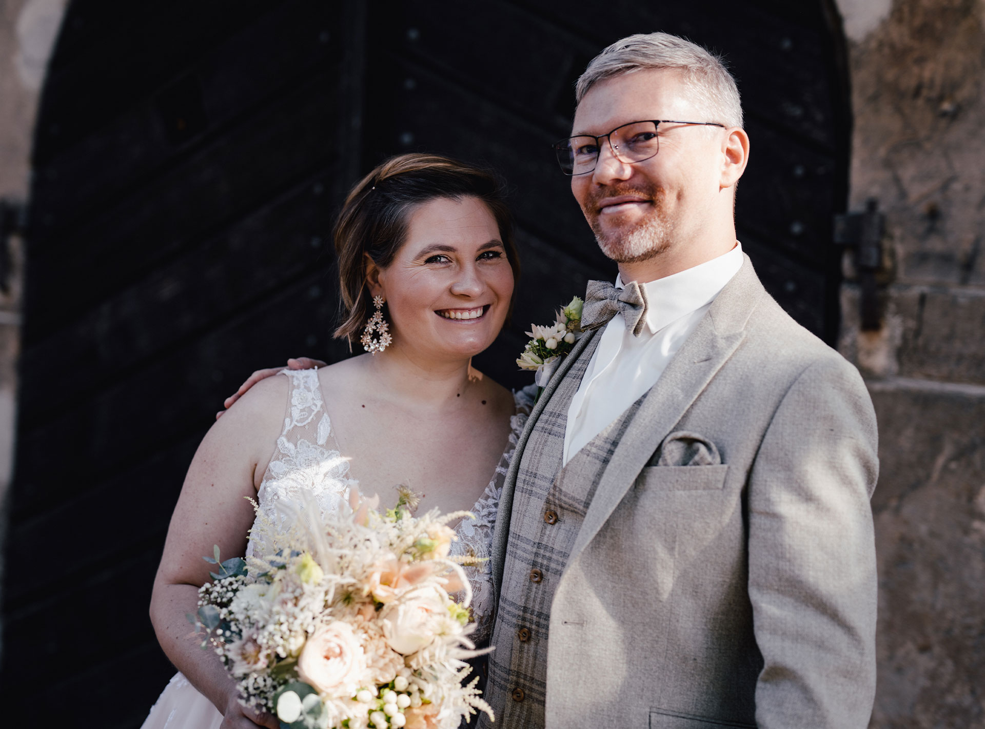 Ein glückliches Brautpaar in eleganter Hochzeitskleidung steht lächelnd vor einer dunklen Holztür, die Braut hält einen Blumenstrauß.
