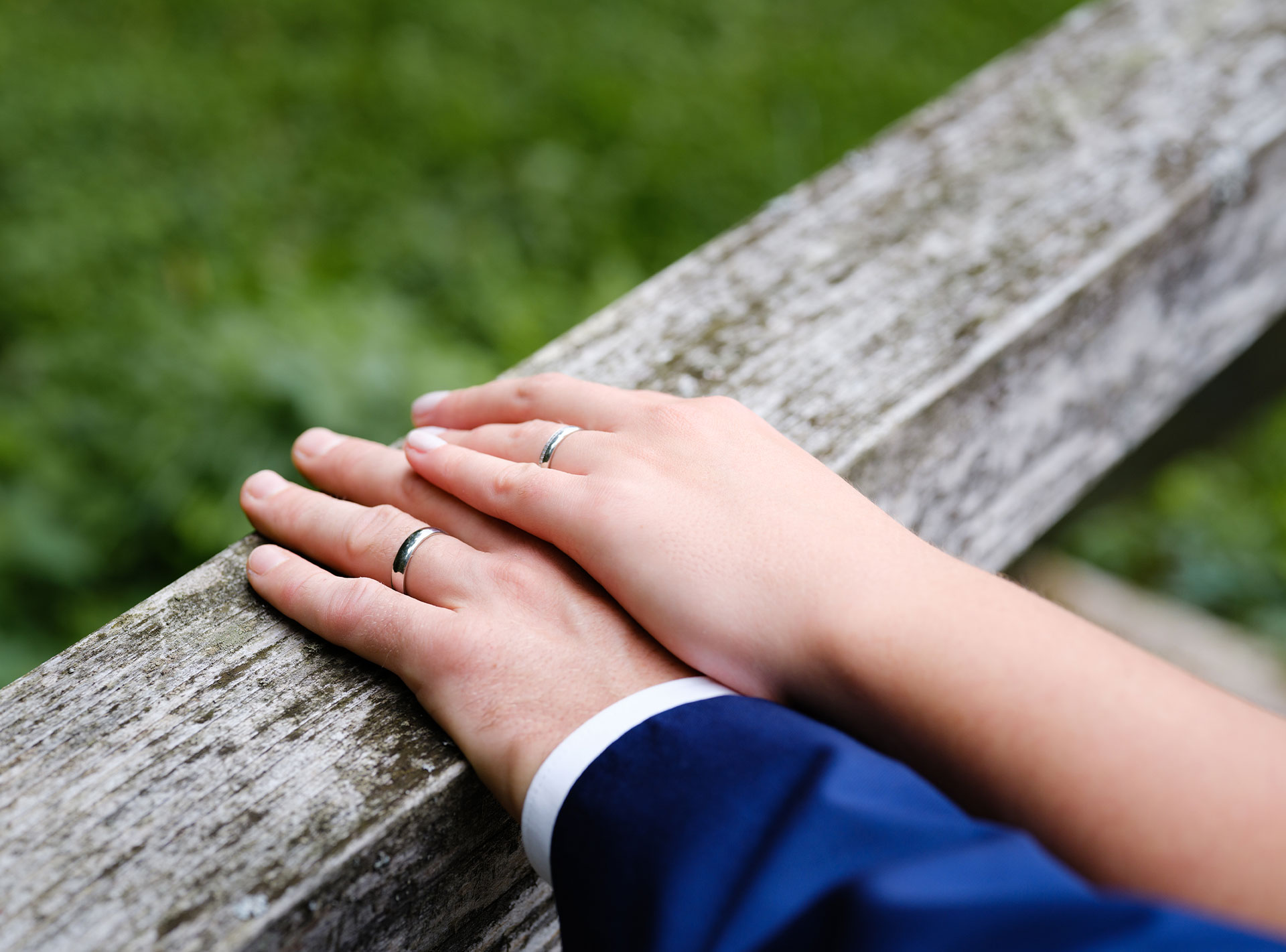 Zwei Hände mit Eheringen auf einem Holzgeländer im Freien.