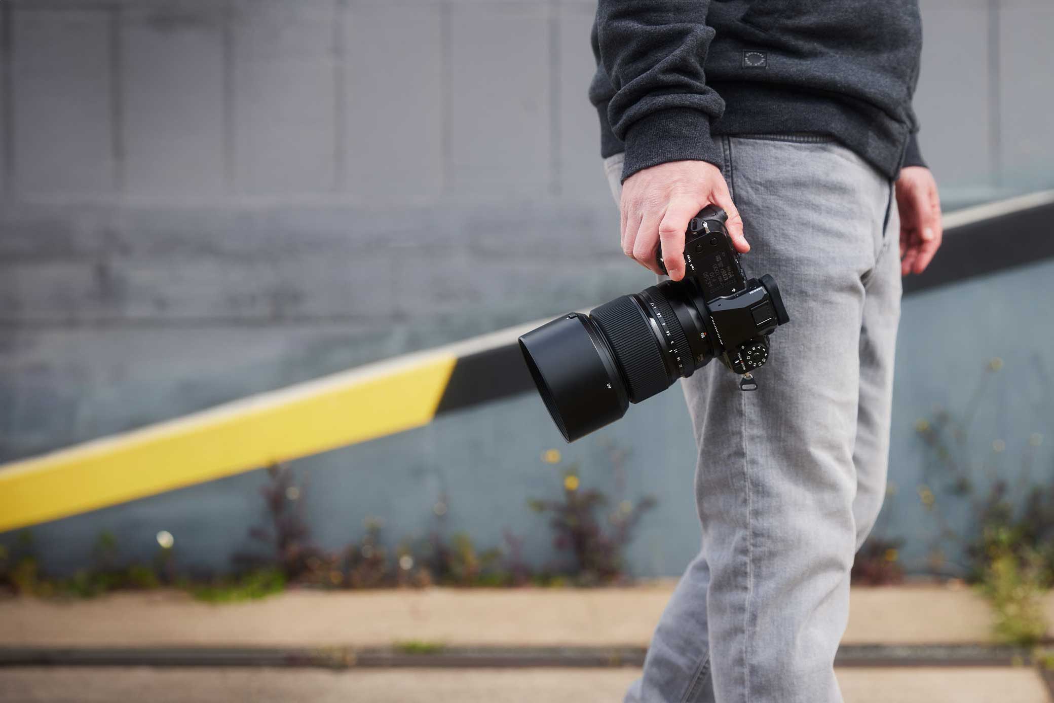 Person hält Kamera mit großem Objektiv in der Hand, graue Hose und Pullover, urbaner Hintergrund.