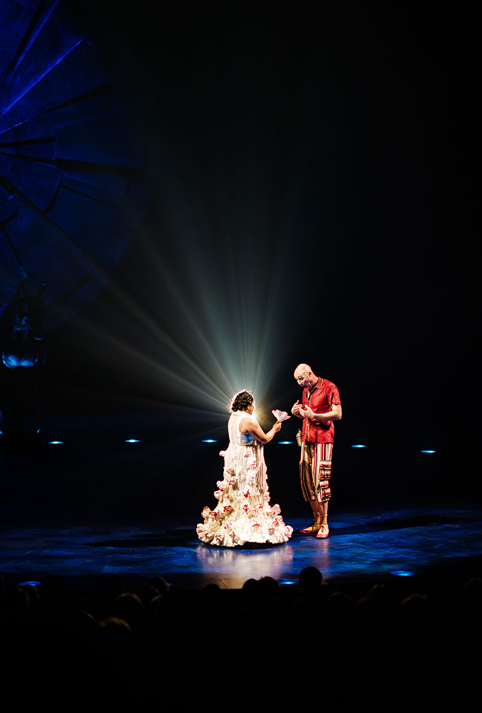 Zwei Schauspieler auf einer Bühne, eine Frau im weißen Kleid mit Blumenmustern und ein Mann in einem bunten Kostüm, im Hintergrund strahlenförmige Lichteffekte.