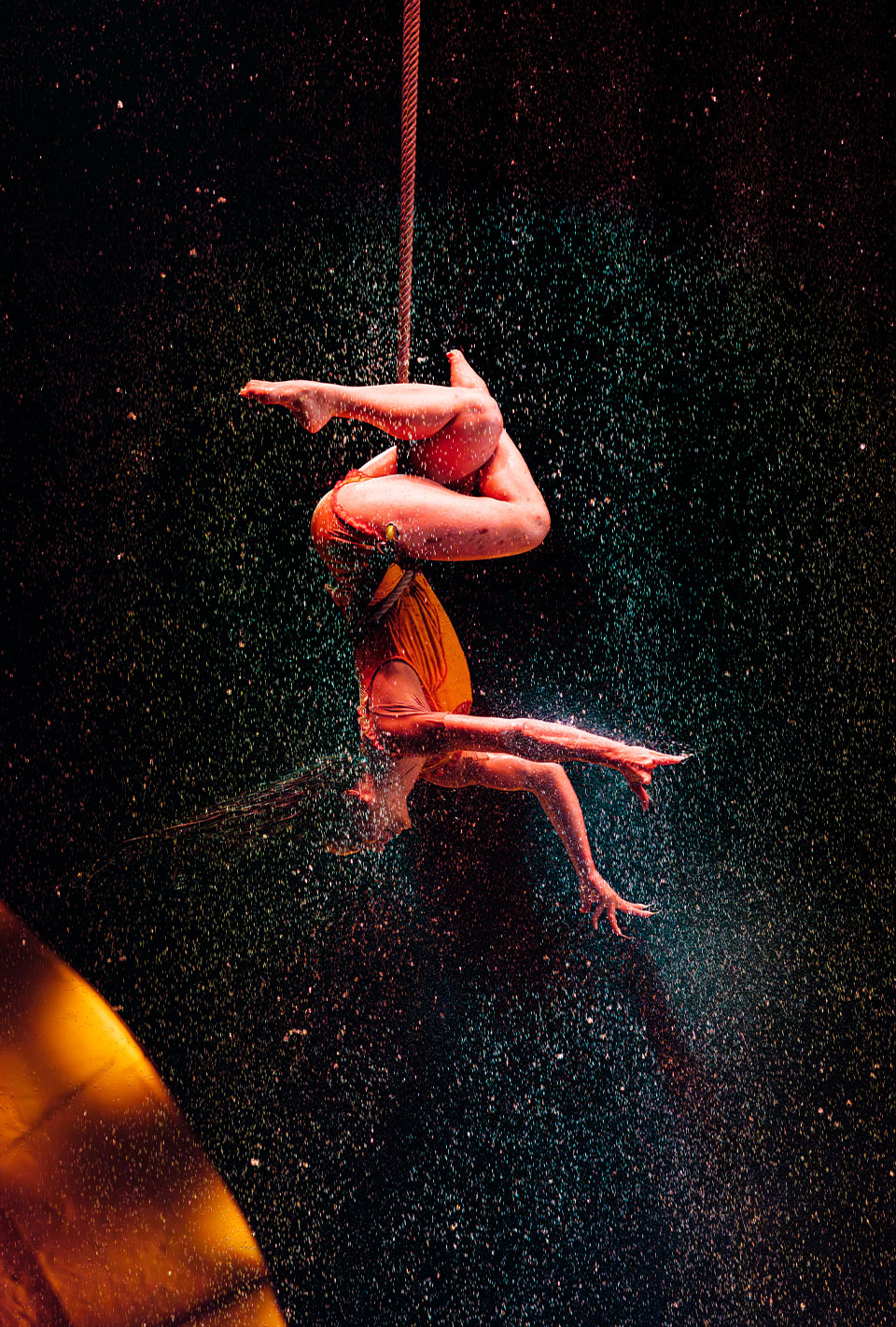 Akrobatin führt eine hängende Figur an einem Seil aus, umgeben von spritzendem Wasser vor einem dunklen Hintergrund.