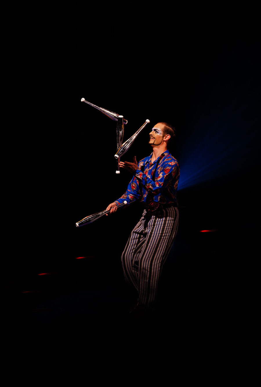 Ein Jongleur mit bemaltem Gesicht und gestreifter Hose jongliert auf der Bühne mit drei Keulen im Scheinwerferlicht.