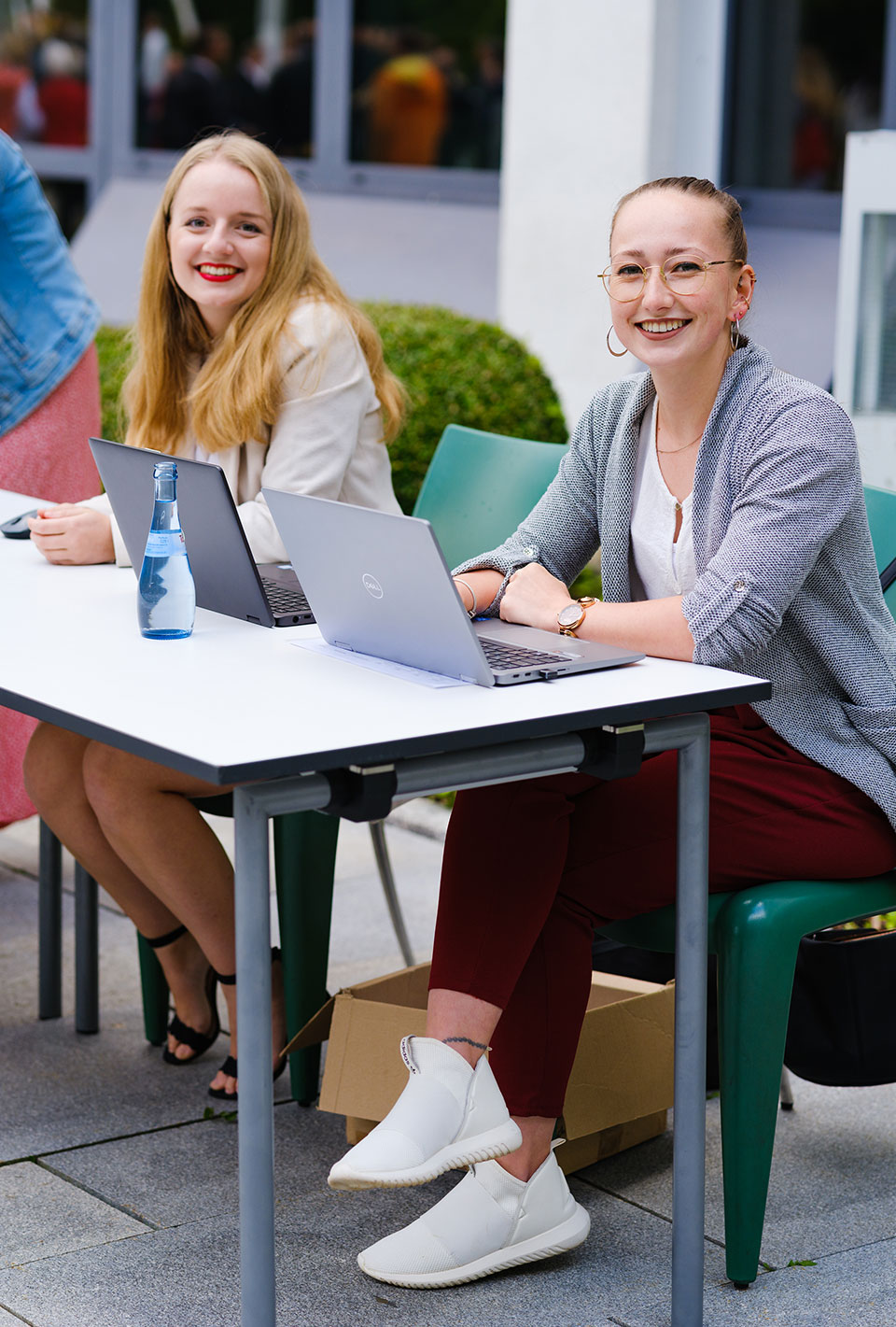 Zwei lächelnde Frauen sitzen draußen an einem Tisch mit Laptops.