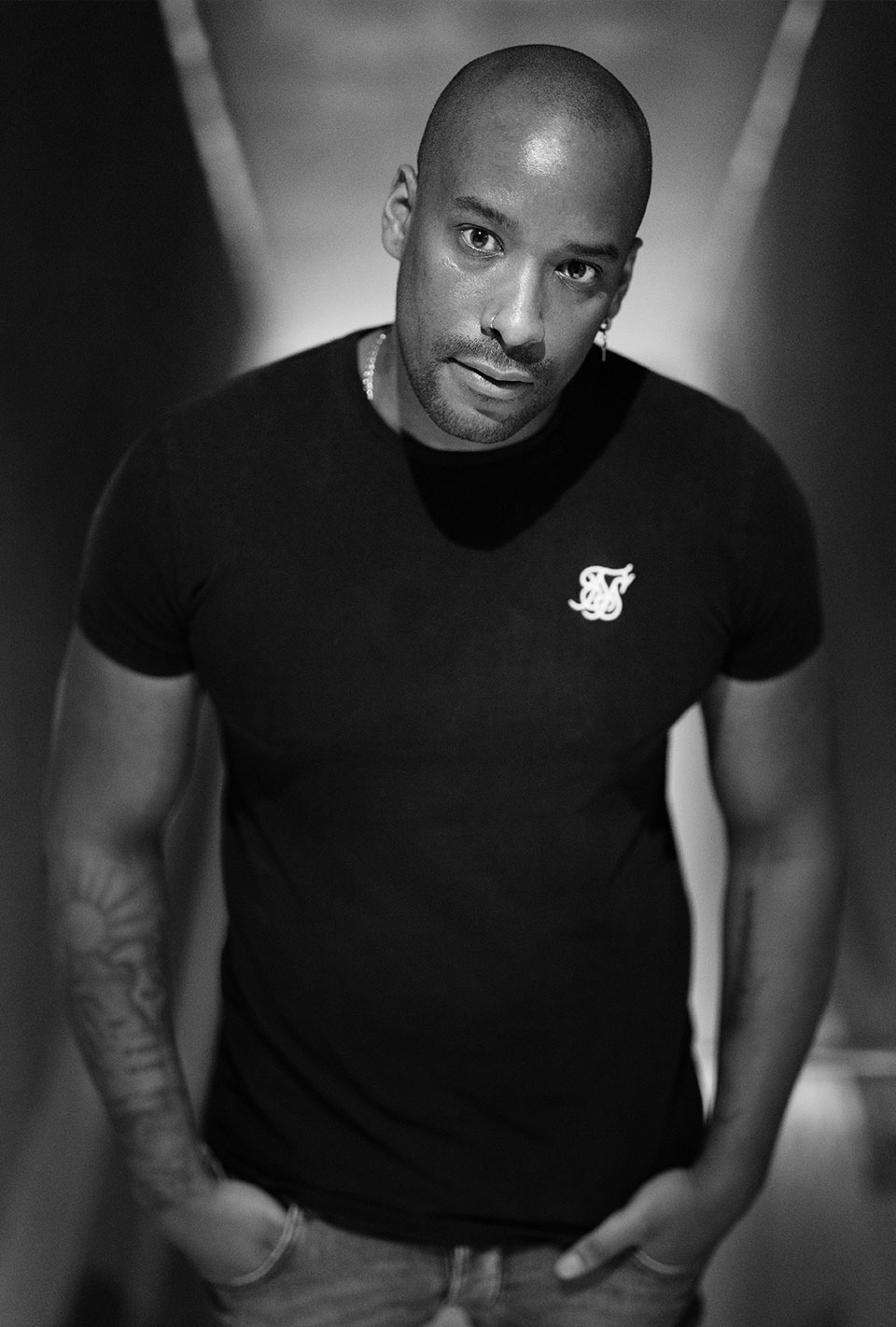 Mann in einem schwarzen T-Shirt, mit den Händen in den Hosentaschen und schaut in die Kamera, Schwarz-Weiß-Fotografie.