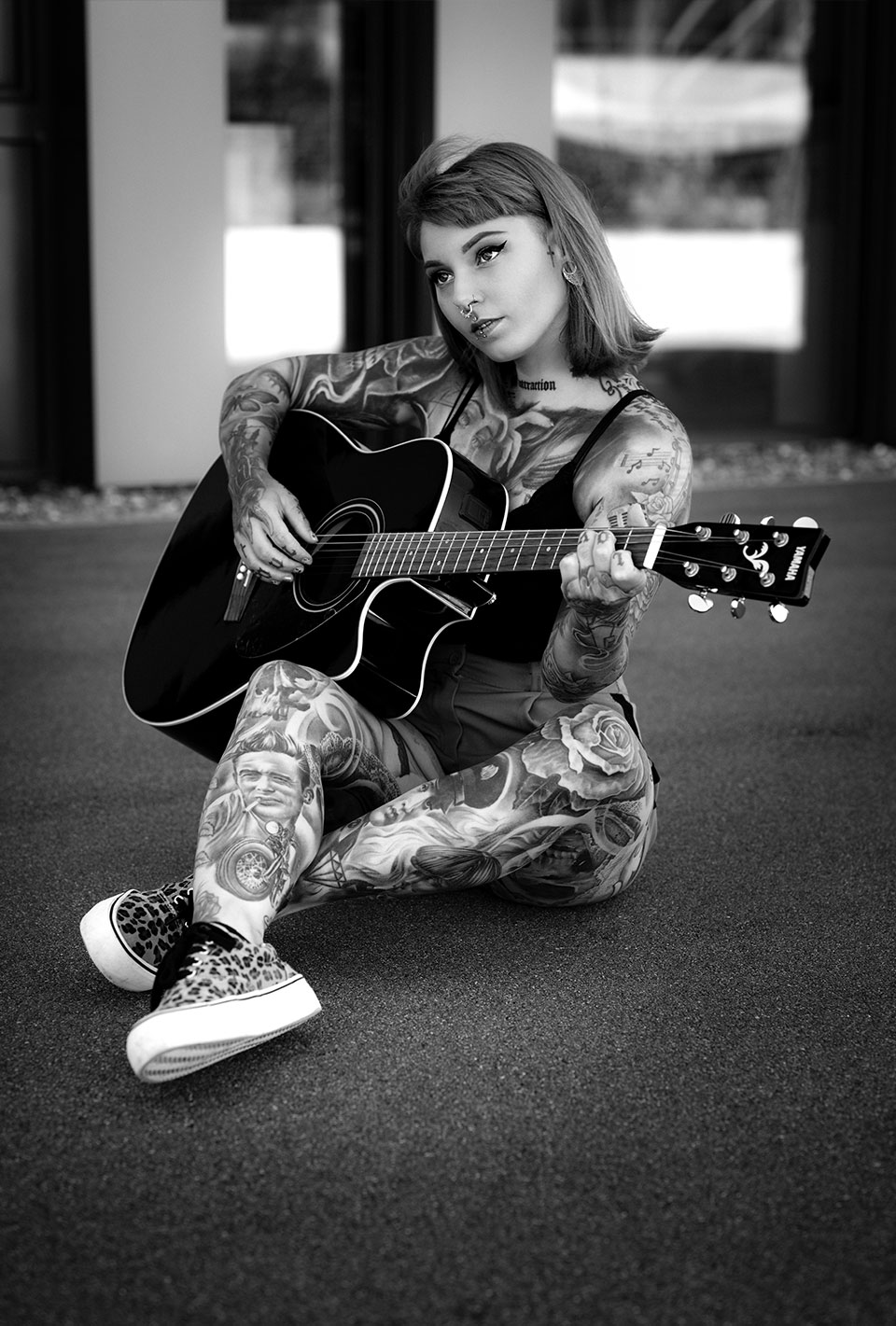 Eine tätowierte Frau mit Piercings sitzt auf dem Boden und spielt Gitarre.