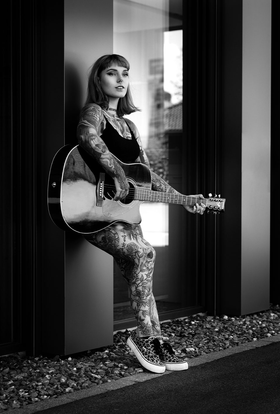 Frau mit Tattoos spielt Gitarre und lehnt an einer Wand.