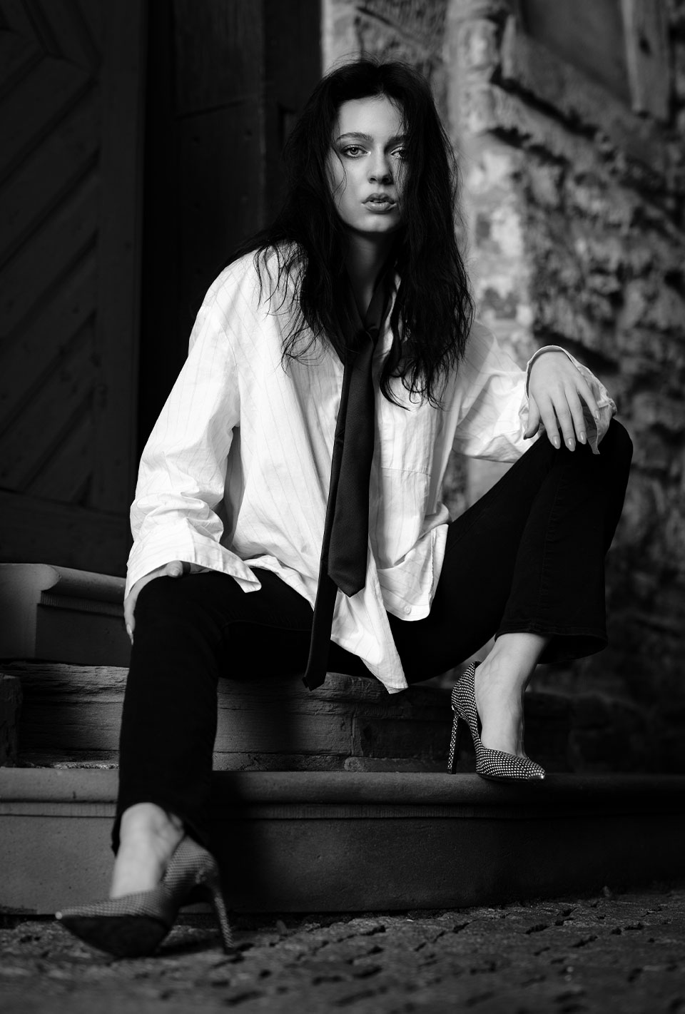 Schwarzweiß-Foto einer Frau mit langen Haaren in einem locker sitzenden Hemd und Krawatte, die auf einer Treppe sitzt und in die Kamera schaut.