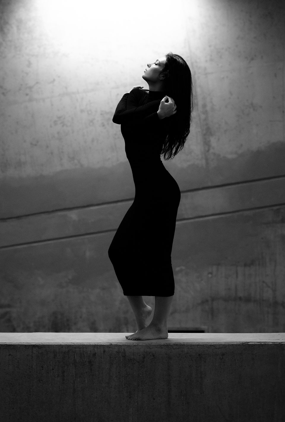 Schwarz-Weiß-Foto einer barfüßigen Frau in einem langen, engen Kleid, die sich in einer Tanzpose vor einer Betonwand bewegt.