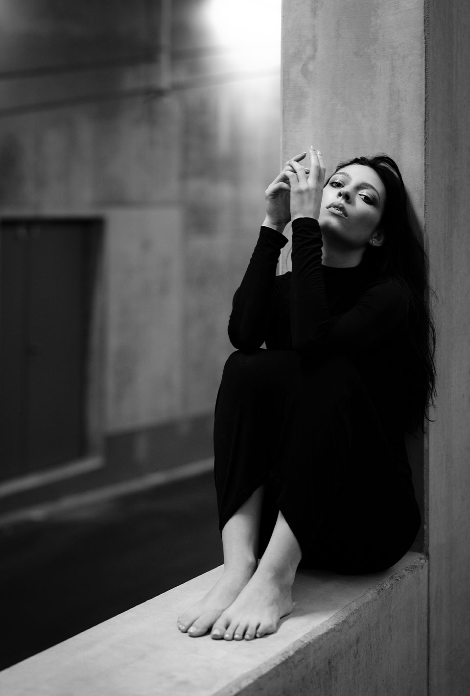 Schwarz-weiße Aufnahme einer Frau in schwarzer Kleidung, die barfuß auf einem Betonvorsprung sitzt und nachdenklich in die Kamera schaut.