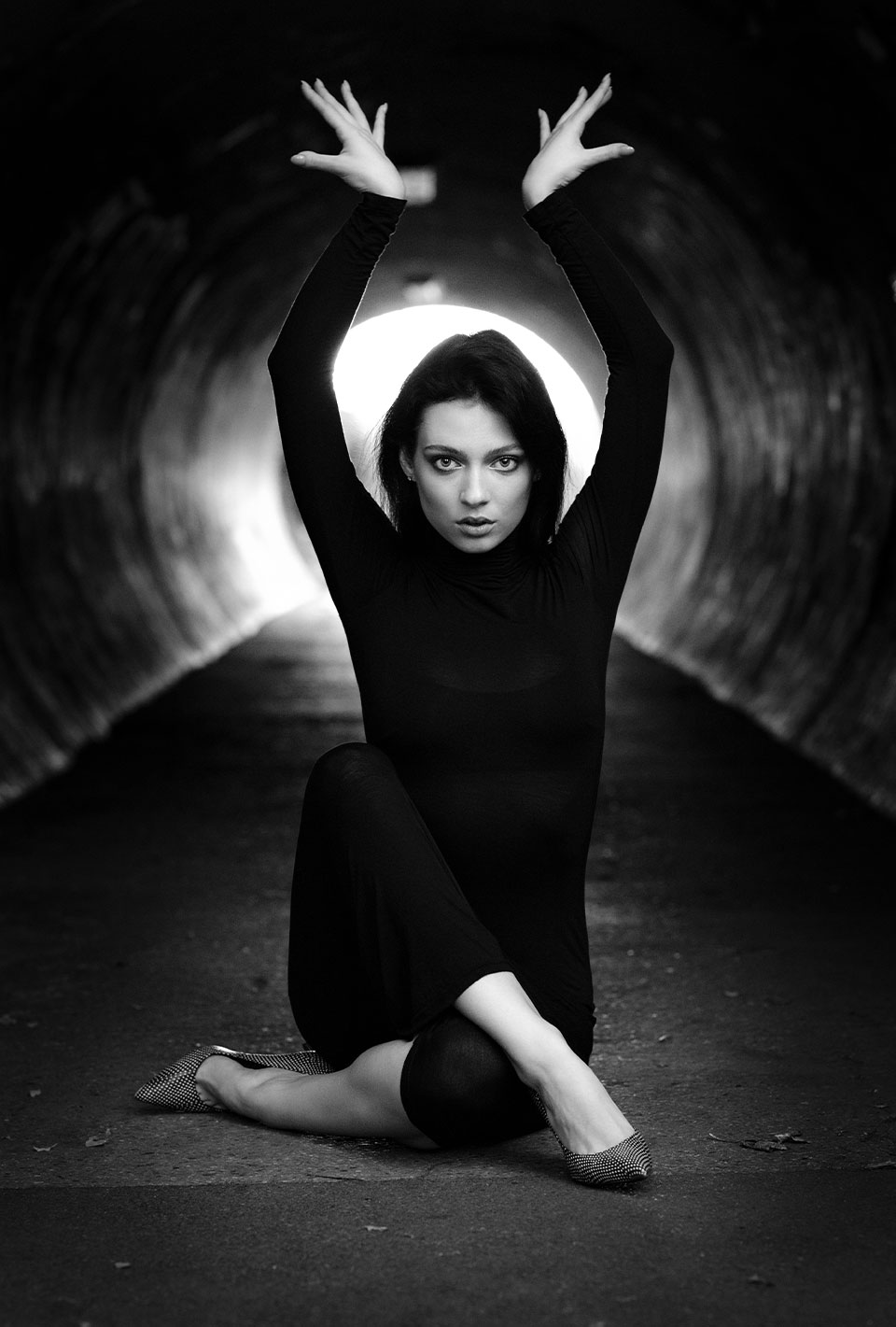 Schwarz-weiß Foto einer Frau in einem langen schwarzen Kleid, die in einem Tunnel in Tanzpose auf dem Boden sitzt und die Arme nach oben streckt.