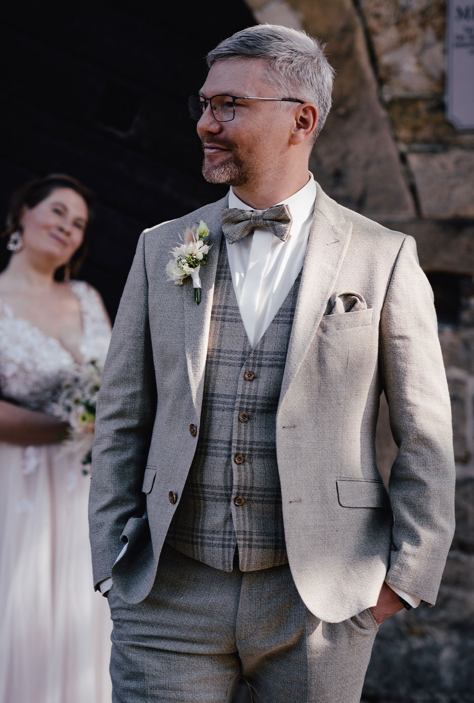 Bräutigam in grauem Anzug und Fliege steht lächelnd, im Hintergrund unscharf die Braut in weißem Kleid.