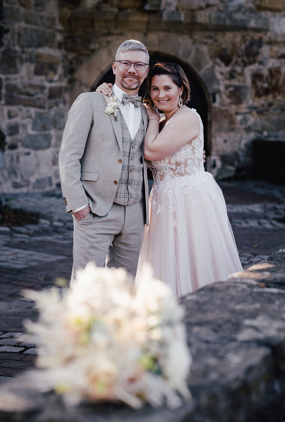 Brautpaar in eleganter Hochzeitskleidung vor einer alten Steinmauer, mit einem unscharfen Blumenstrauß im Vordergrund.