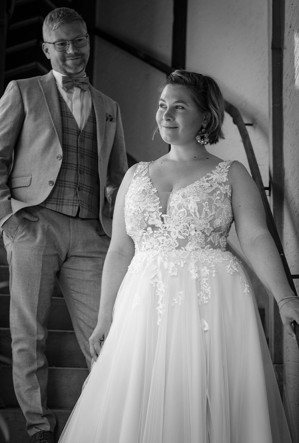 Braut und Bräutigam stehen auf einer Treppe, die Braut trägt ein Spitzenhochzeitskleid und der Bräutigam einen Anzug mit Weste und Fliege.