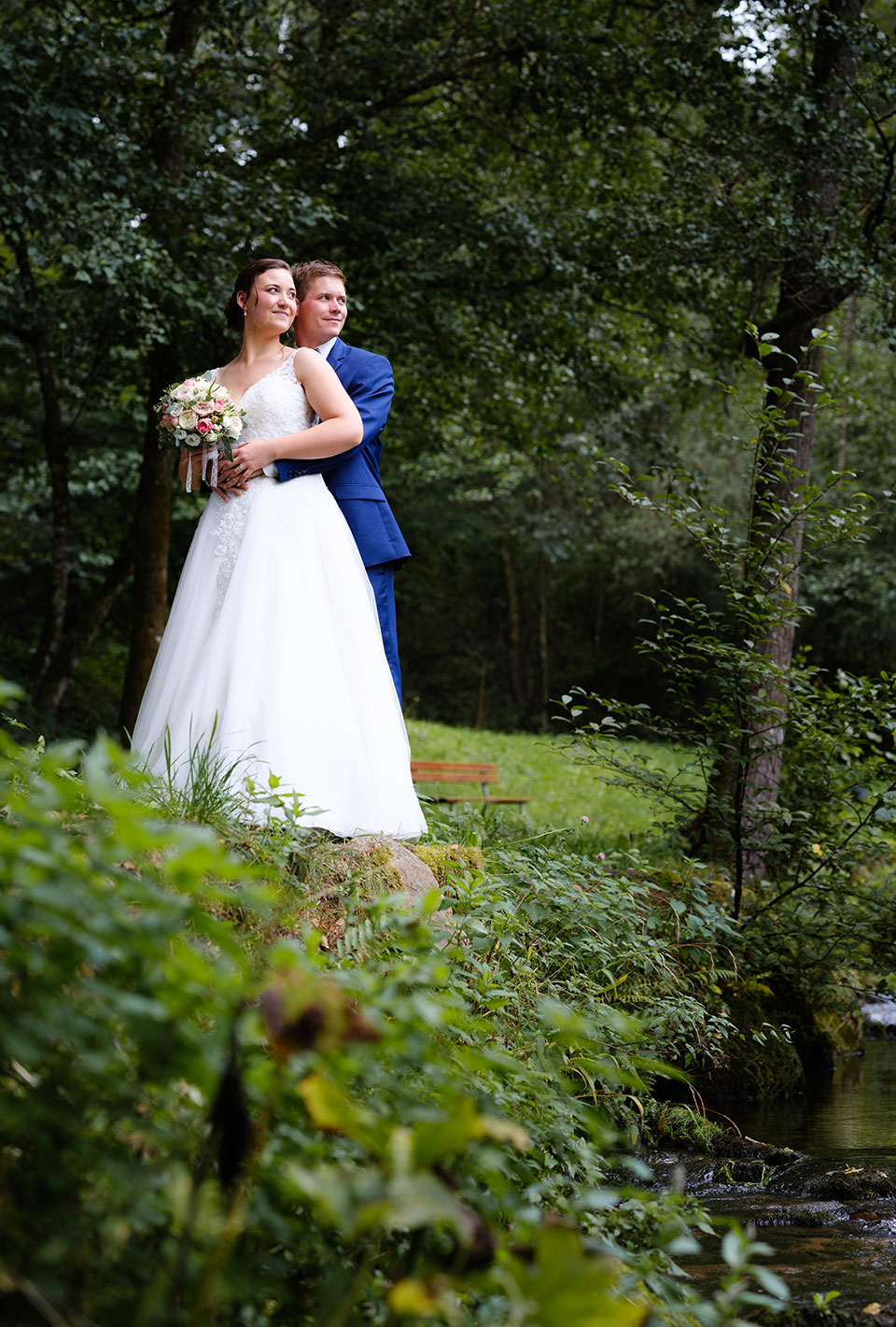 Brautpaar im Hochzeitskleid und Anzug posiert im Grünen, umgeben von Bäumen und Natur.