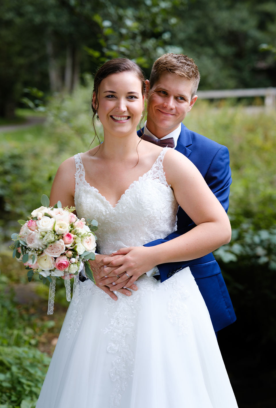 Brautpaar steht lächelnd im Freien, Braut hält Blumenstrauß.