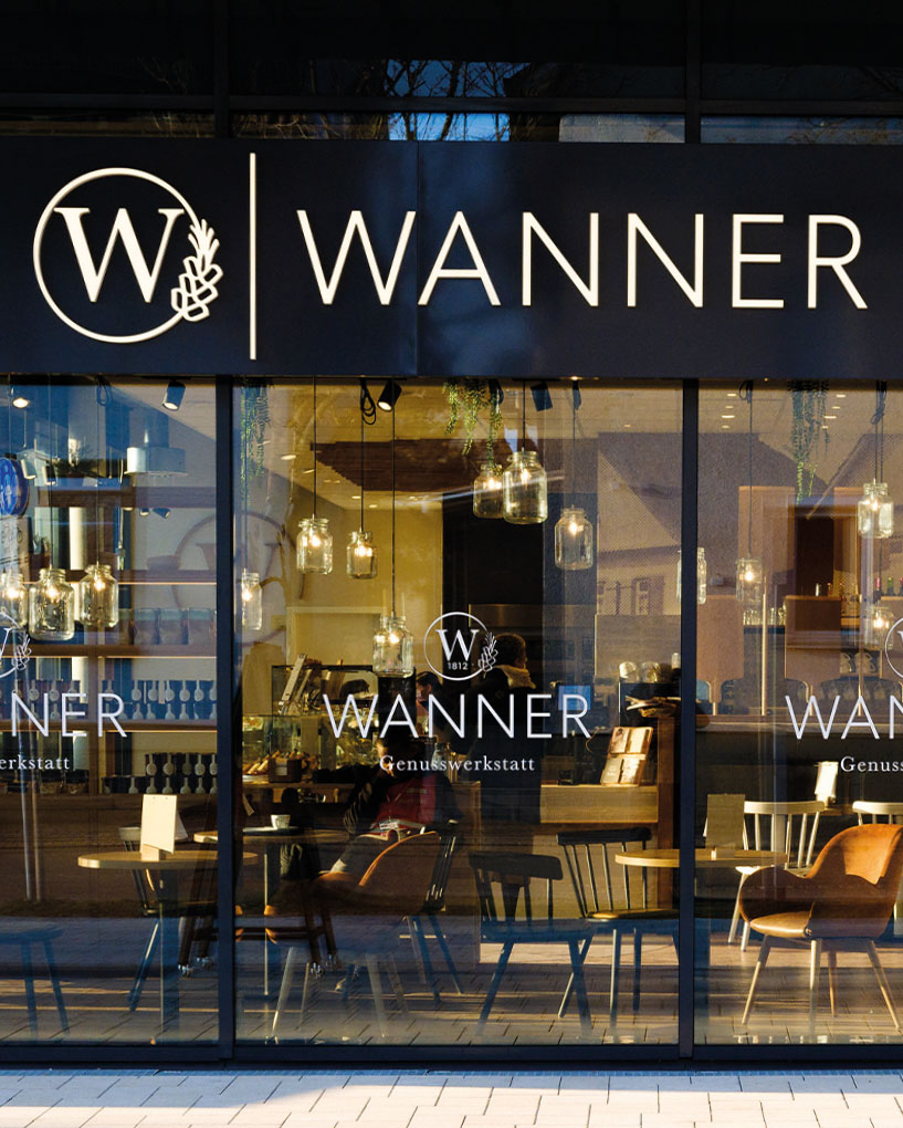 Außenansicht eines modernen Cafés mit dem Namen Wanner, große Fensterfront, schlichte Holz- und Metallstühle, hängende Glühlampen im Innenraum.